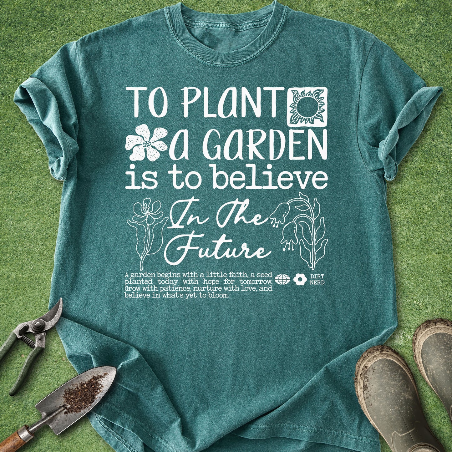 A teal short-sleeve graphic T-shirt laid flat on green grass-like background, with gardening tools and boots around it. The shirt has large white text reading 'TO PLANT A GARDEN IS TO BELIEVE In The Future' with flower and sun illustrations.