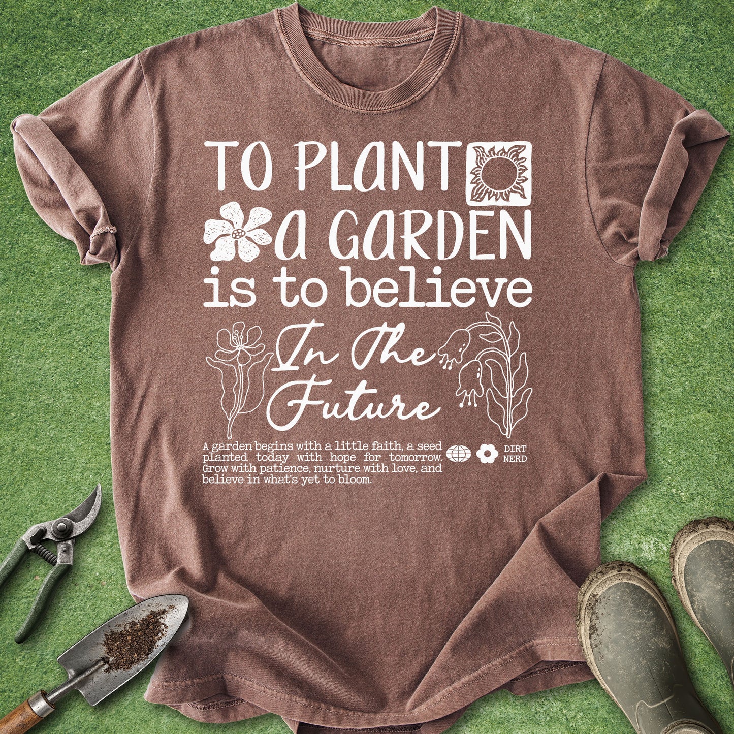 A brown short-sleeve graphic T-shirt laid flat on green grass-like background, with gardening tools and boots around it. The shirt has white text reading 'TO PLANT A GARDEN IS TO BELIEVE In The Future' with small floral and plant illustrations.
