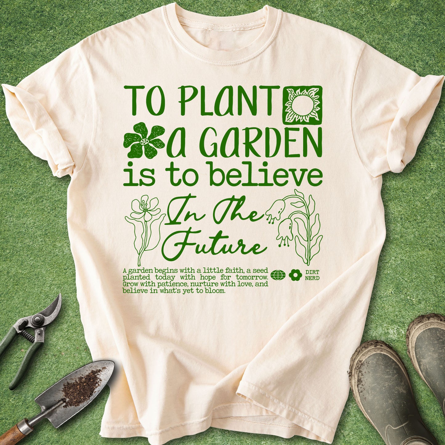A cream-colored short-sleeve t-shirt with a green graphic print that reads 'TO PLANT A GARDEN IS TO BELIEVE In The Future' accompanied by gardening illustrations. The shirt is laid flat on a green grass-like surface with gardening tools (trowel, shears) and shoes as props.
