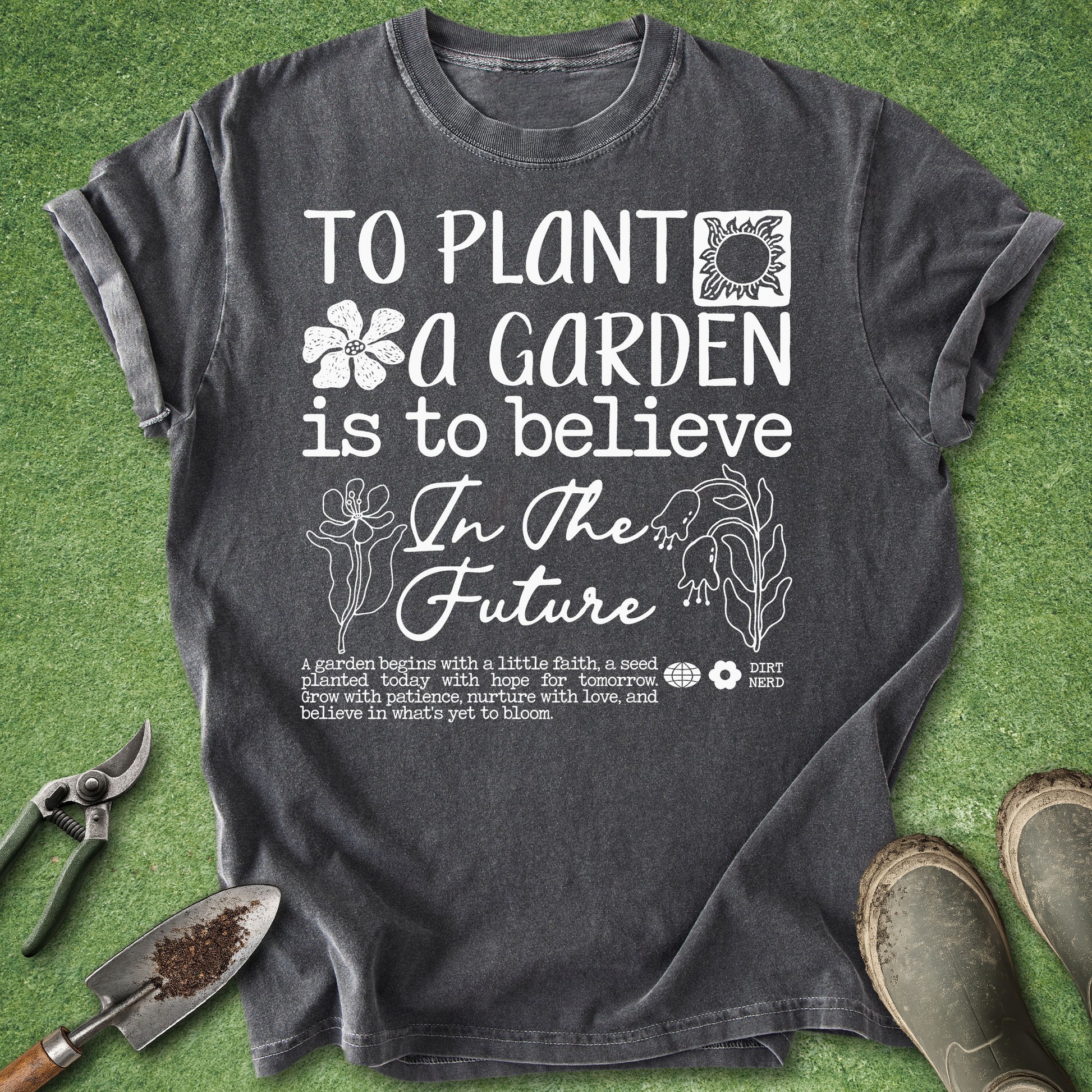 A dark gray graphic t-shirt laid flat on green grass with gardening props including pruning shears, a trowel, and gardening boots. The shirt features a white printed design with the text 'TO PLANT A GARDEN IS TO BELIEVE In The Future' and botanical illustrations.