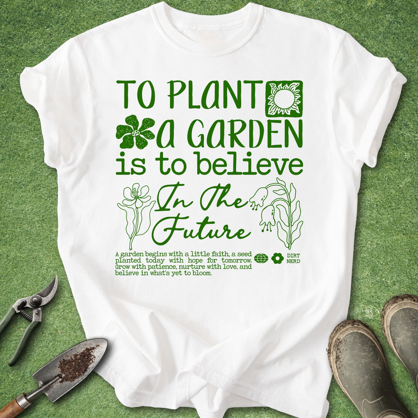 A white short-sleeve graphic T-shirt laid flat on green grass-like background, with gardening tools and shoes around it. The shirt has green text reading 'TO PLANT A GARDEN is to believe In The Future' with flower and leaf illustrations.