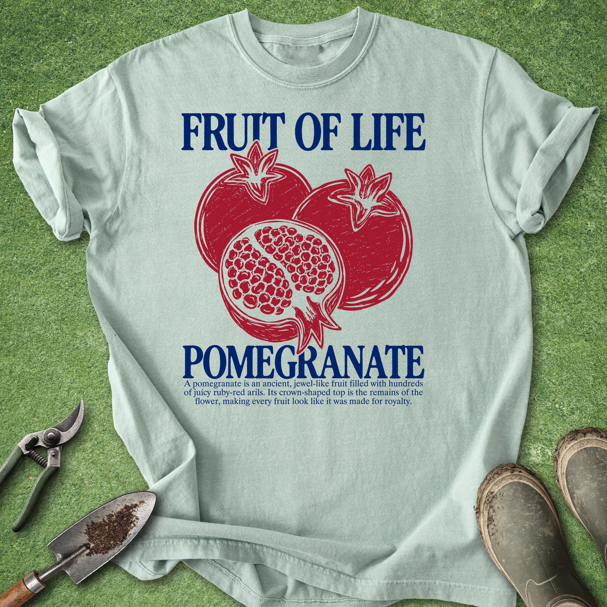 A light blue graphic t-shirt laid flat on a green grass-like surface, featuring a red pomegranate illustration and the words 'FRUIT OF LIFE' and 'POMEGRANATE' in blue. Gardening tools and work boots are placed around the shirt as props.