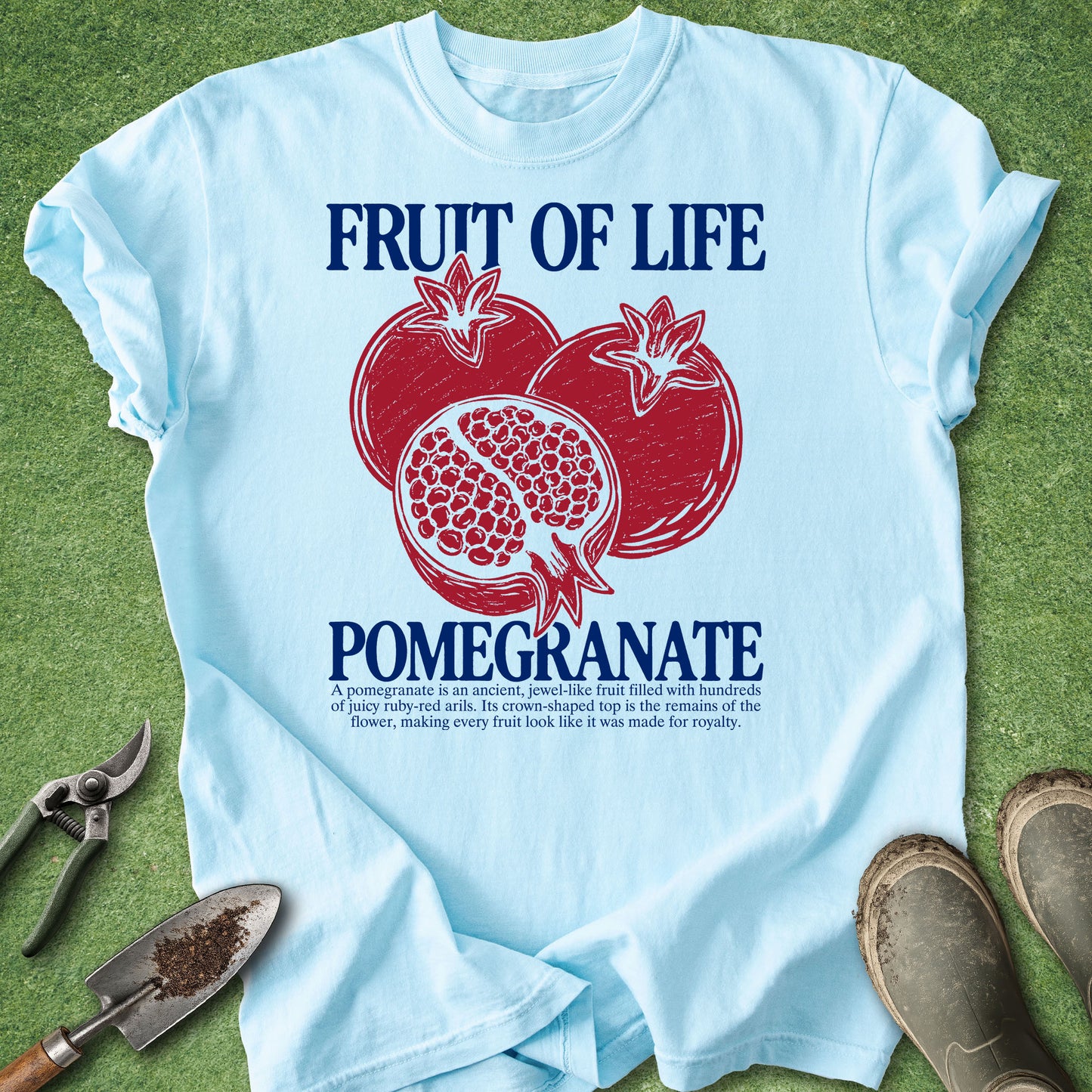 A light blue graphic t-shirt laid flat on green grass, featuring a red pomegranate illustration and the text 'FRUIT OF LIFE POMEGRANATE' in dark blue. Gardening props including pruning shears, a trowel, and boots are visible around the shirt.