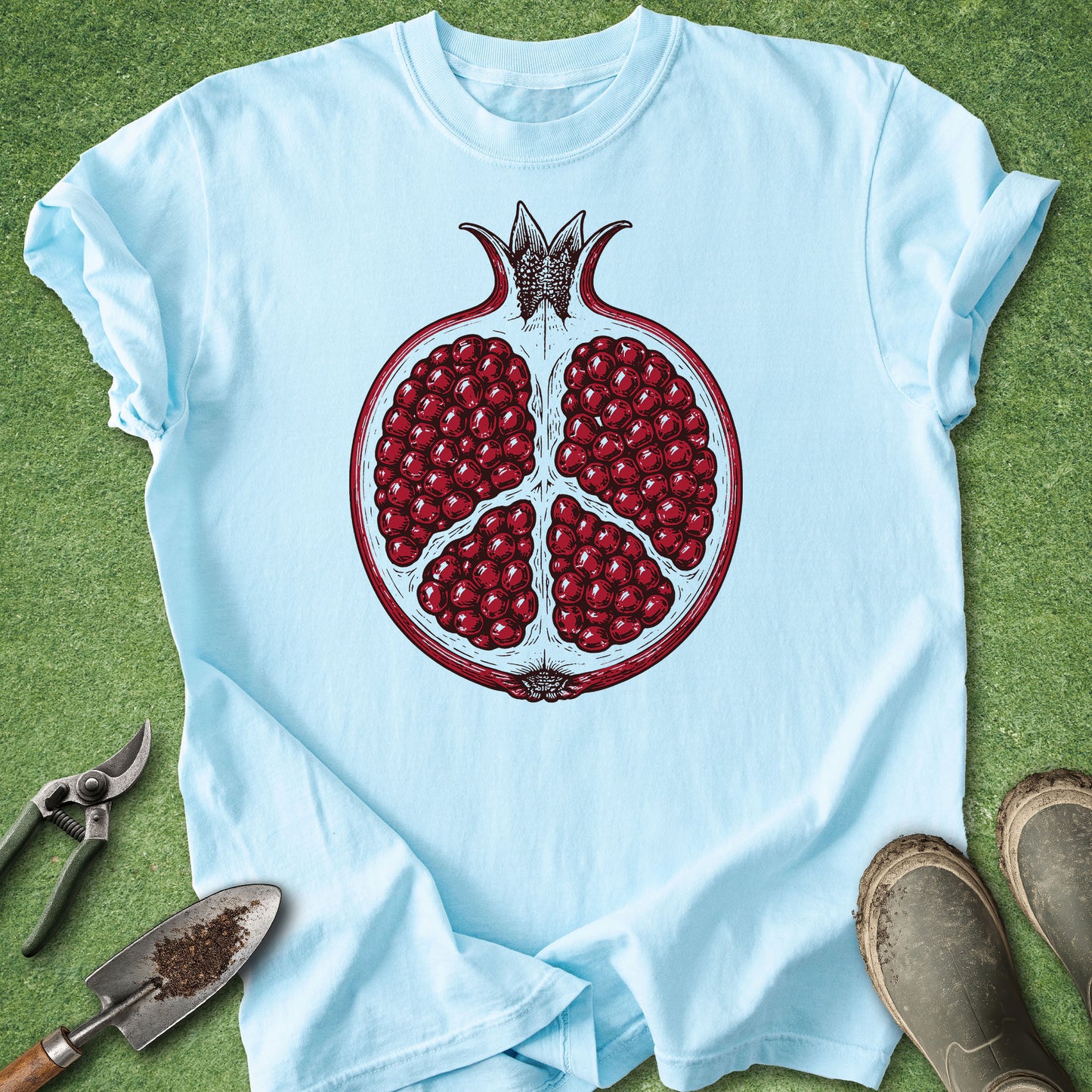A light blue short-sleeve t-shirt laid flat on green grass, featuring a large graphic of a cut-open pomegranate with red seeds on the front. Gardening tools and brown boots are placed around the shirt as styled props.