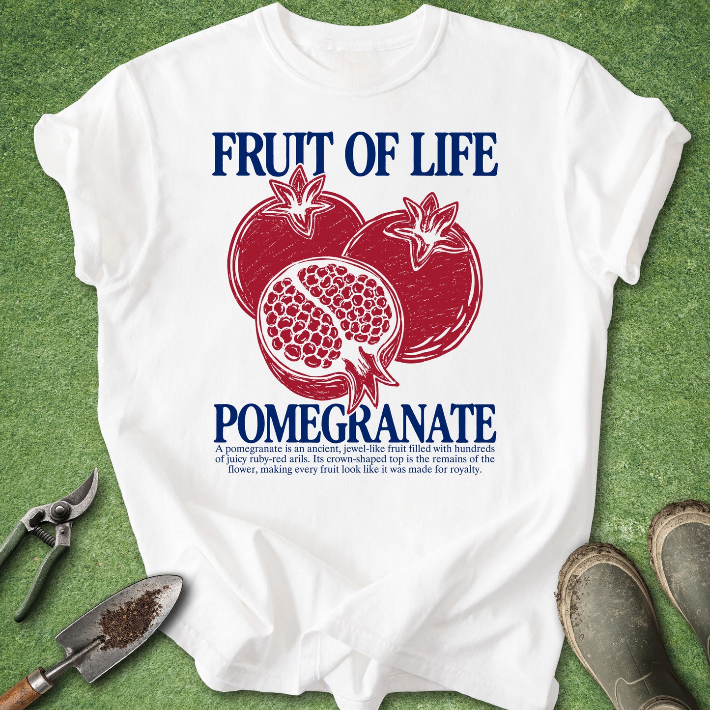 A white short-sleeve graphic T-shirt laid flat on a green grass-like surface, featuring a red pomegranate illustration and blue text reading “FRUIT OF LIFE POMEGRANATE,” with small descriptive text below. Gardening props including pruning shears, a trowel, and work boots are visible around the shirt.