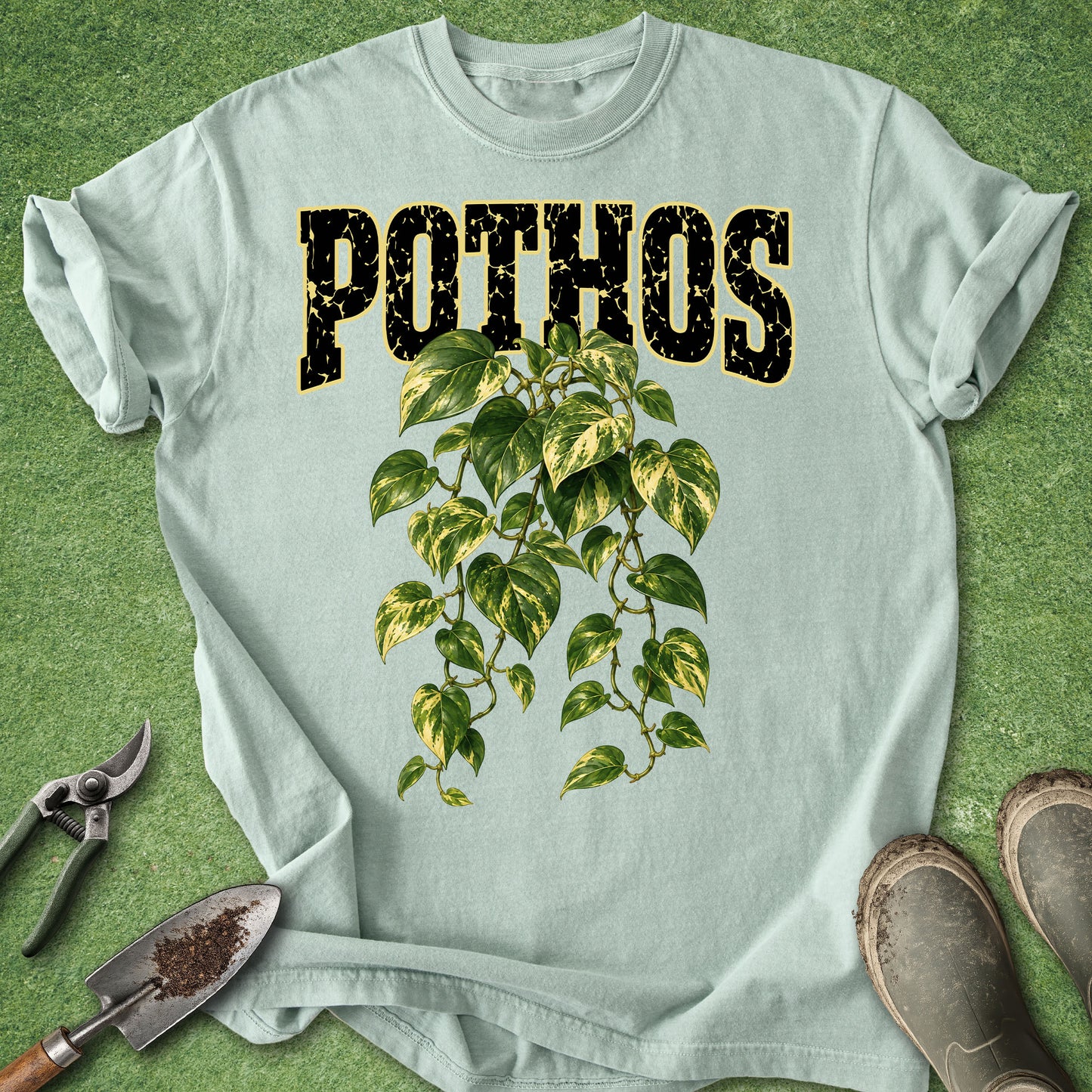 A light blue crewneck t-shirt laid flat on a green grass-like surface, featuring the word 'POTHOS' in distressed black letters outlined in gold above a green ivy plant graphic. Gardening tools and boots are placed around the shirt as styling props.