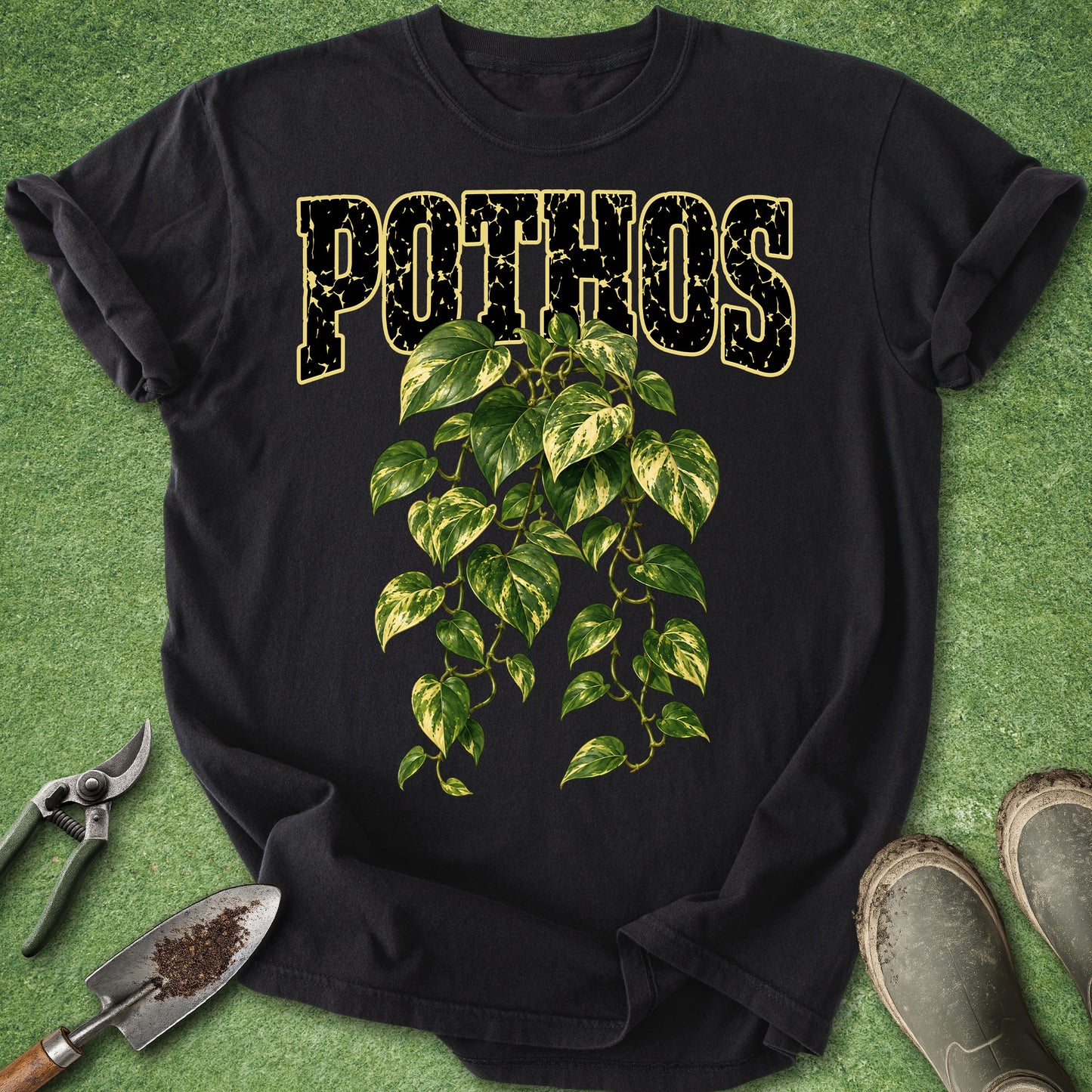A black short-sleeve graphic T-shirt laid flat on a green grass-like surface, with the word “POTHOS” in distressed gold letters above a green and yellow leafy vine illustration. Gardening tools and muddy boots are placed around the shirt as styling props.