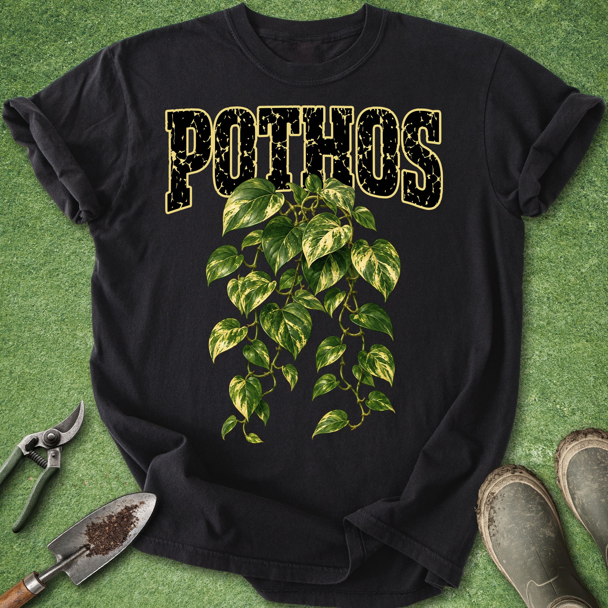 A black short-sleeve graphic T-shirt laid flat on a green grass-like surface, with the word “POTHOS” in distressed gold letters above a green and yellow leafy vine illustration. Gardening tools and muddy boots are placed around the shirt as styling props.