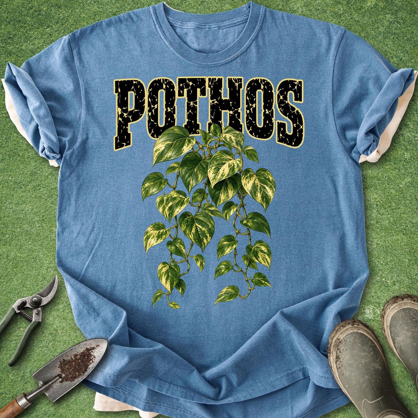 A blue short-sleeve T-shirt laid flat on a green grass-like surface, featuring the word 'POTHOS' in distressed black letters outlined in gold above a green leafy vine graphic. Gardening props including pruning shears, a small trowel, and boots are placed around the shirt.