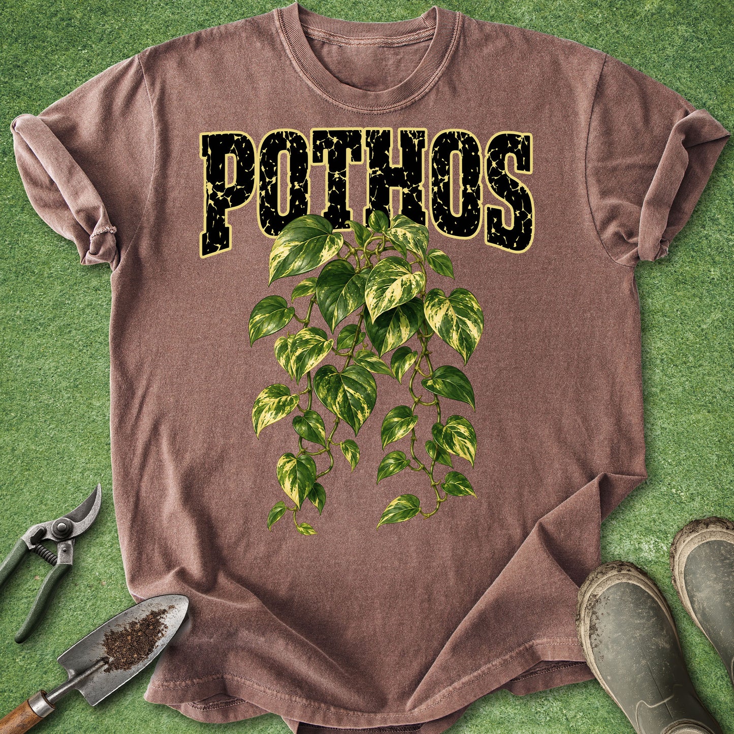 A brown short-sleeve T-shirt laid flat on green artificial grass, featuring the word 'POTHOS' in distressed black letters outlined in yellow above a green and cream variegated leafy vine graphic. Gardening props including pruning shears, a small hand trowel, and muddy boots are placed around the shirt.