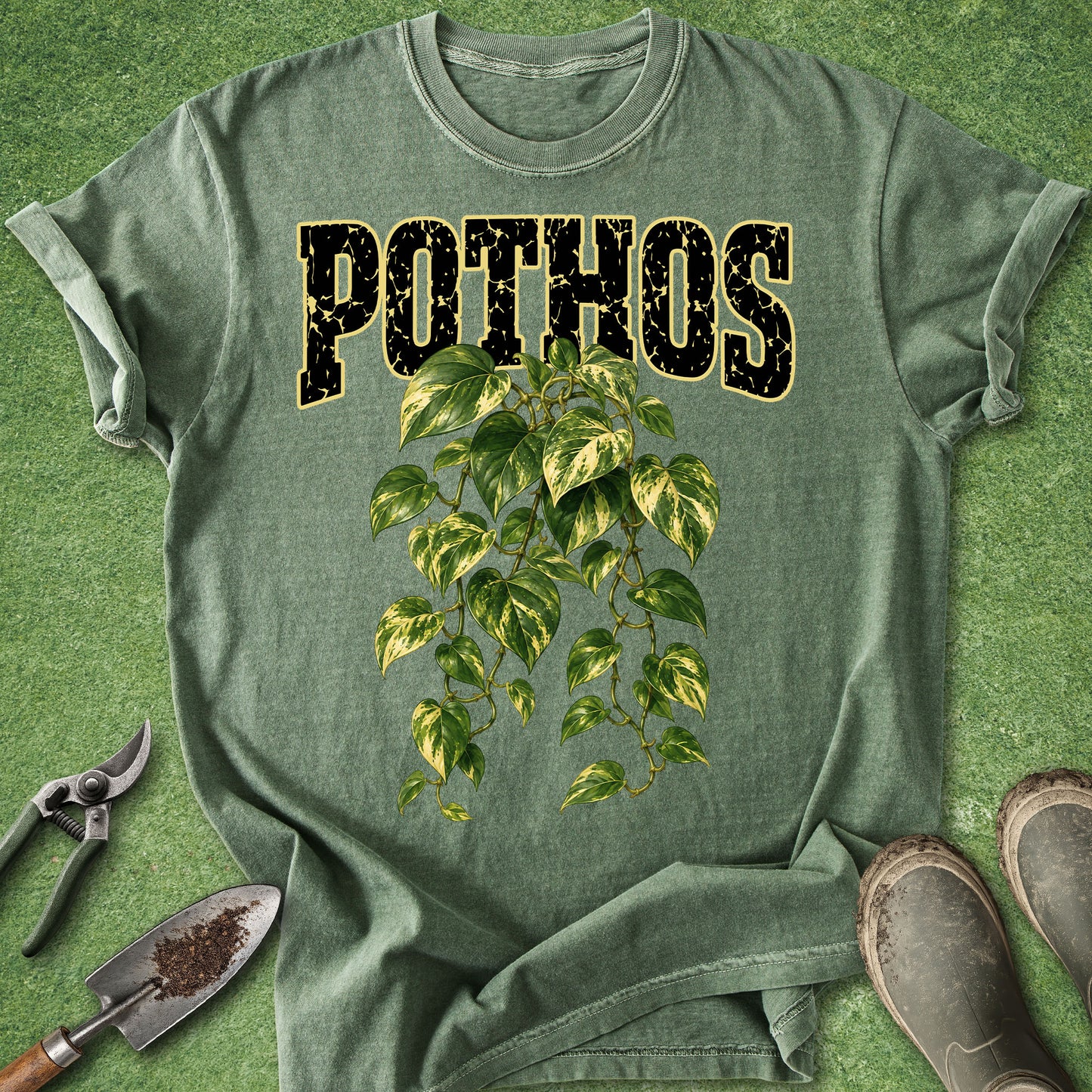 A sage green t-shirt laid flat on a green grass-like surface, featuring the word 'POTHOS' in distressed black letters outlined in gold above a green and yellow variegated leafy vine graphic. Gardening props including a trowel, pruning shears, and boots are arranged around the shirt.