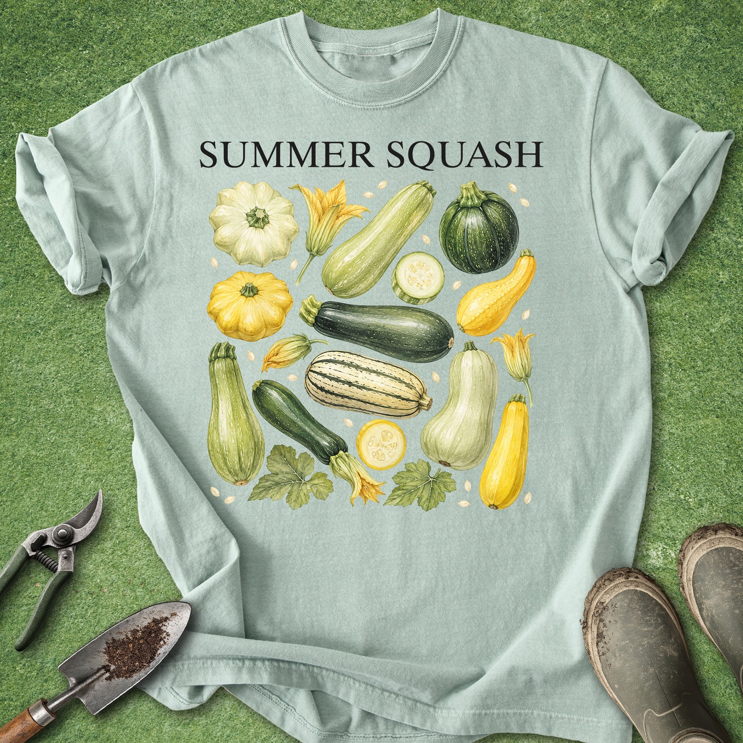 A light blue-green graphic t-shirt laid flat on a green grass-like surface, featuring the words 'SUMMER SQUASH' above an illustration of various vegetables including zucchini, yellow squash, corn, and melon. Gardening tools and boots are placed around the shirt as styling props.