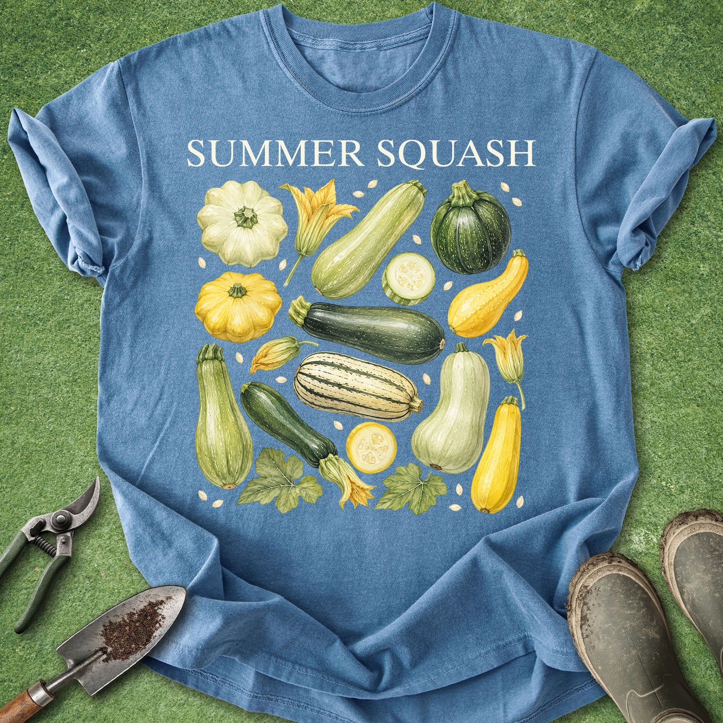 A blue short-sleeve T-shirt laid flat on a green grass-like surface, printed with the words “SUMMER SQUASH” above an illustrated arrangement of various squash and zucchini vegetables. Gardening props including pruning shears, a small hand trowel, and muddy boots are placed around the shirt.