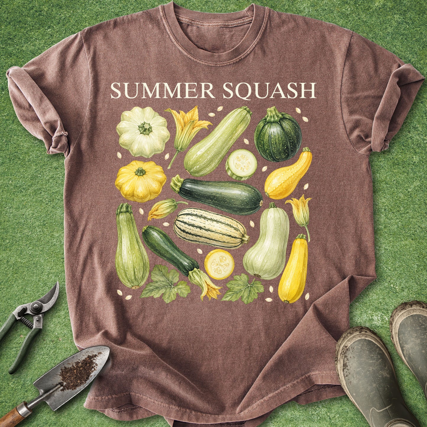 A brown short-sleeve graphic T-shirt laid flat on green grass-like ground, with the words "SUMMER SQUASH" above an illustrated assortment of vegetables including zucchini, cucumber, squash, melon, and pumpkin. Gardening tools and boots are partially visible around the shirt.