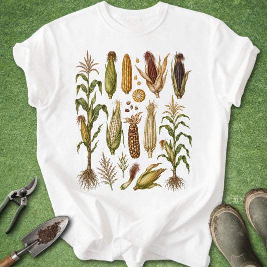 A white short-sleeve T-shirt laid flat on a green textured surface, featuring a vintage-style botanical illustration of various corn cobs, ears, seeds, and plant details printed on the front. Gardening tools and muddy boots are placed around the shirt as styling props.
