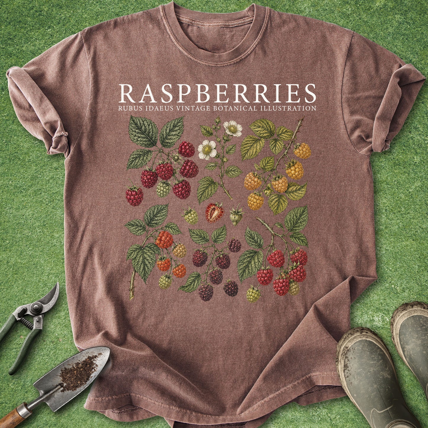 A heathered brown short-sleeve t-shirt laid flat on green grass, featuring a vintage-style botanical illustration of raspberries and blackberries with the text 'RASPBERRIES' and 'RUBUS IDAEUS VINTAGE BOTANICAL ILLUSTRATION'. Gardening tools and boots are placed around the shirt as props.