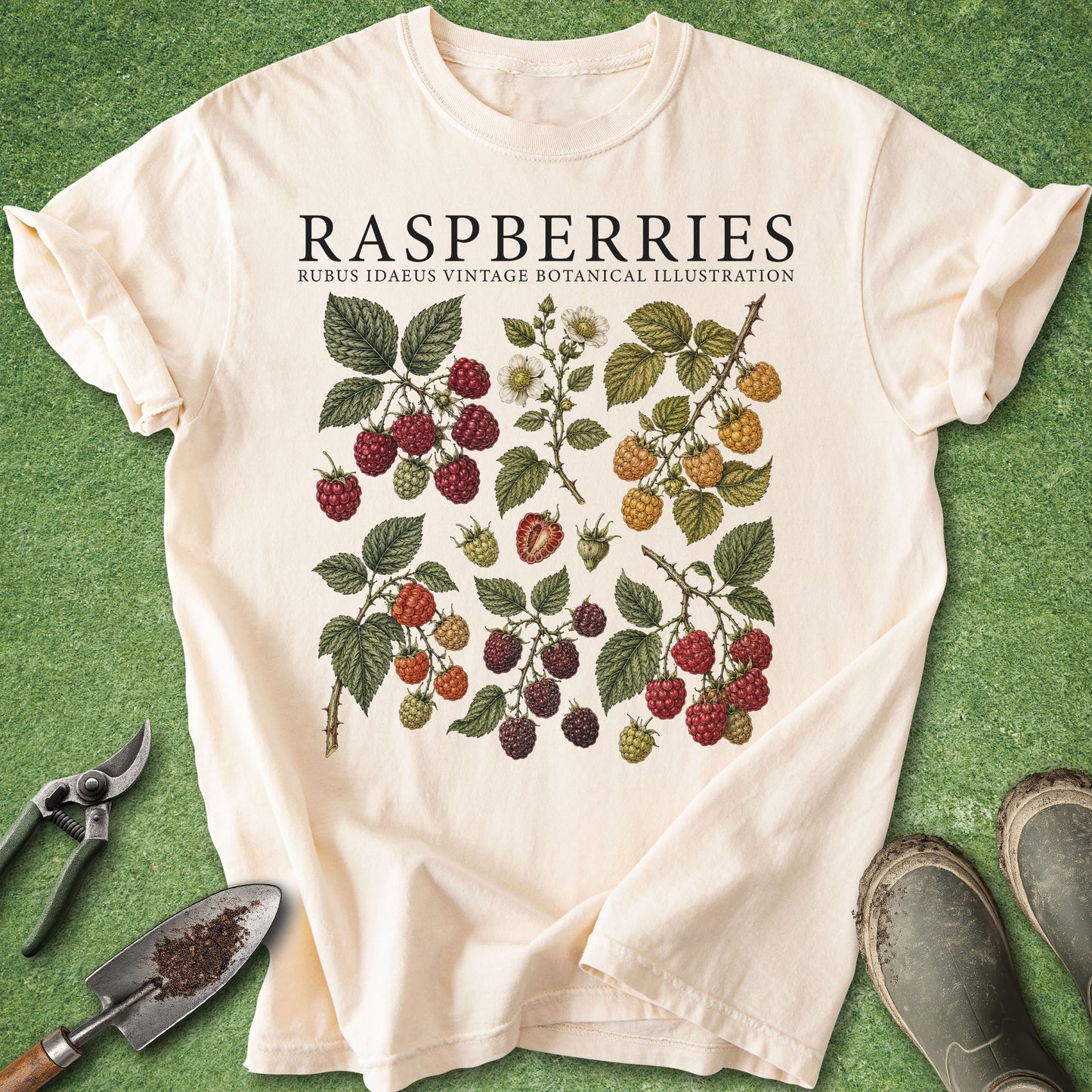 An off-white short-sleeve t-shirt with a vintage botanical illustration of raspberries, laid flat on a green grass-like surface with gardening tools and boots nearby.
