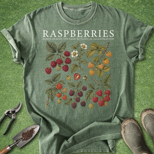 A sage green graphic t-shirt laid flat on a green grass-like surface, featuring a vintage-style raspberry botanical illustration and the text 'RASPBERRIES RUBUS IDAEUS VINTAGE BOTANICAL ILLUSTRATION.' Gardening props including pruning shears, a trowel, and boots are placed around the shirt.