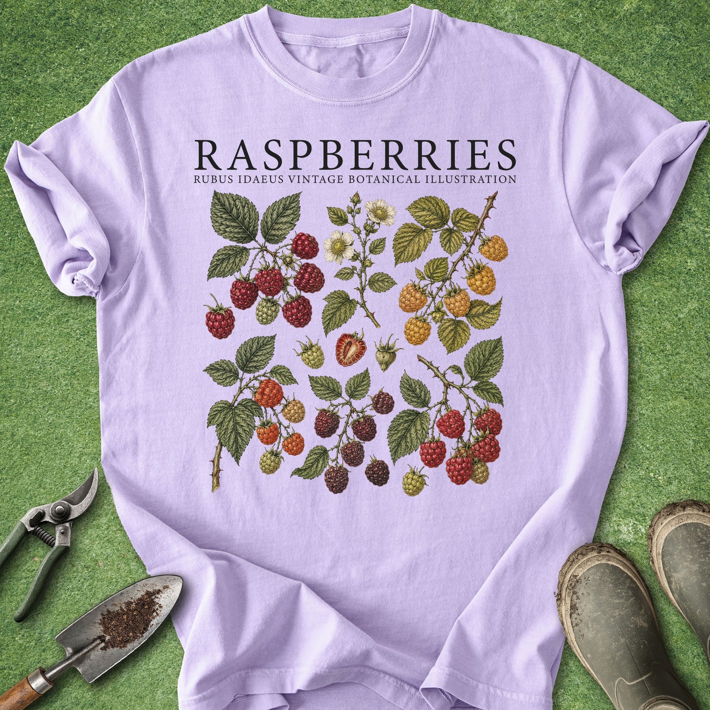 A lavender crewneck t-shirt with a vintage-style botanical illustration of raspberries on a green grass-like background, styled with gardening tools and boots.
