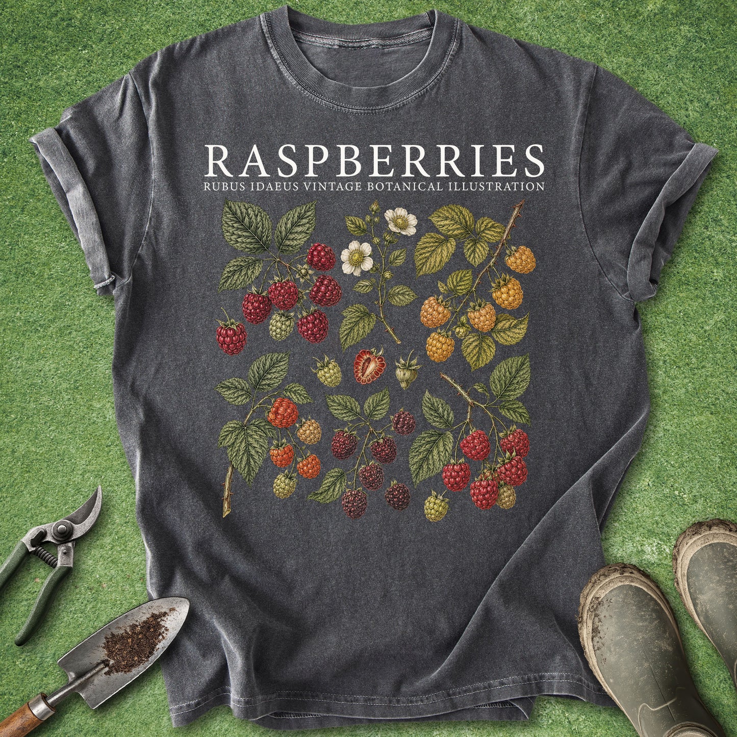 A dark gray graphic t-shirt featuring a vintage botanical illustration of raspberries and blackberries with the text 'RASPBERRIES RUBUS IDAEUS VINTAGE BOTANICAL ILLUSTRATION'. The shirt is styled in a flat-lay on green grass with gardening tools (trowel, pruning shears) and muddy boots.