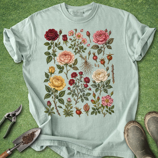 A light mint green t-shirt with a vintage botanical illustration of roses and berries, laid flat on a green grass-like surface with gardening tools and boots nearby.