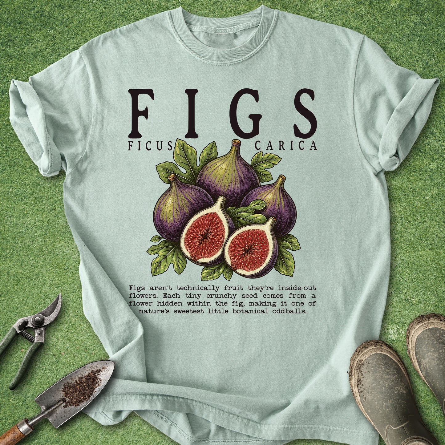 A light mint green graphic t-shirt laid flat on a green grass-like surface, featuring a detailed illustration of four figs (one whole, three halves) with leaves and the text 'FIGS FICUS CARICA' above. Gardening props like a trowel, pruning shears, and boots are visible around the shirt.