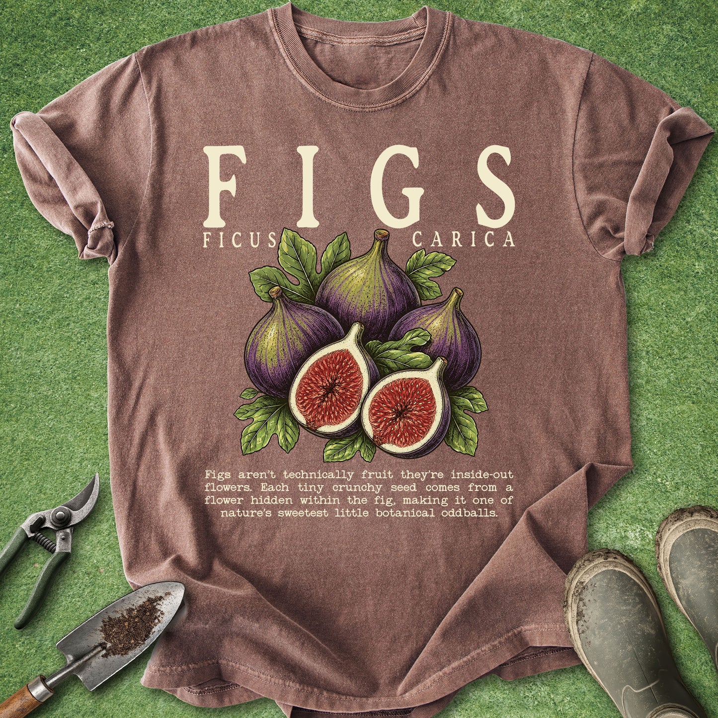 A brown graphic t-shirt laid flat on green grass-like background, featuring an illustrated figs design with the text 'FIGS FICUS CARICA' and a small botanical fact. Gardening props including pruning shears, a small hand trowel, and boots are placed around the shirt.