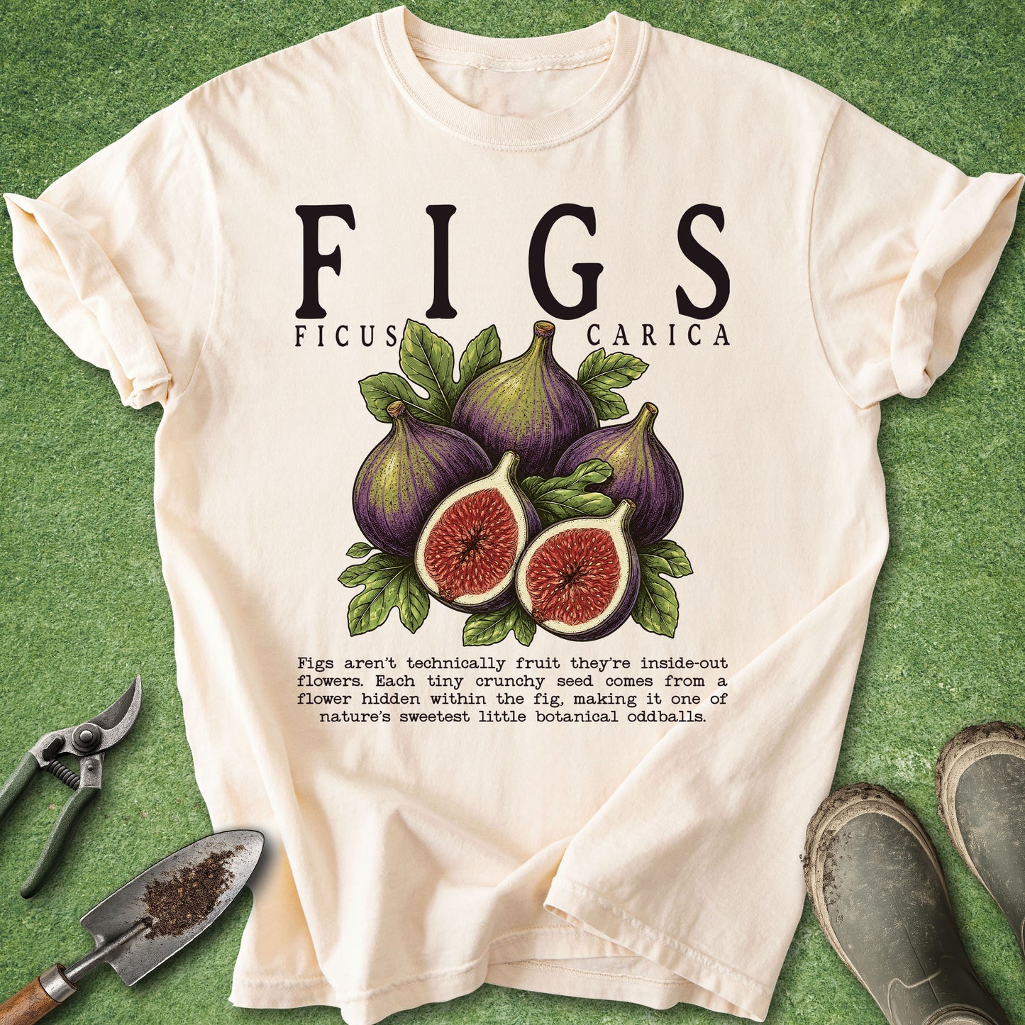 A cream-colored graphic t-shirt featuring an illustration of purple figs and the text 'FIGS FICUS CARICA' with a botanical fact. The shirt is laid flat on a green grass-like surface, styled with gardening tools (trowel, pruning shears) and a pair of boots.