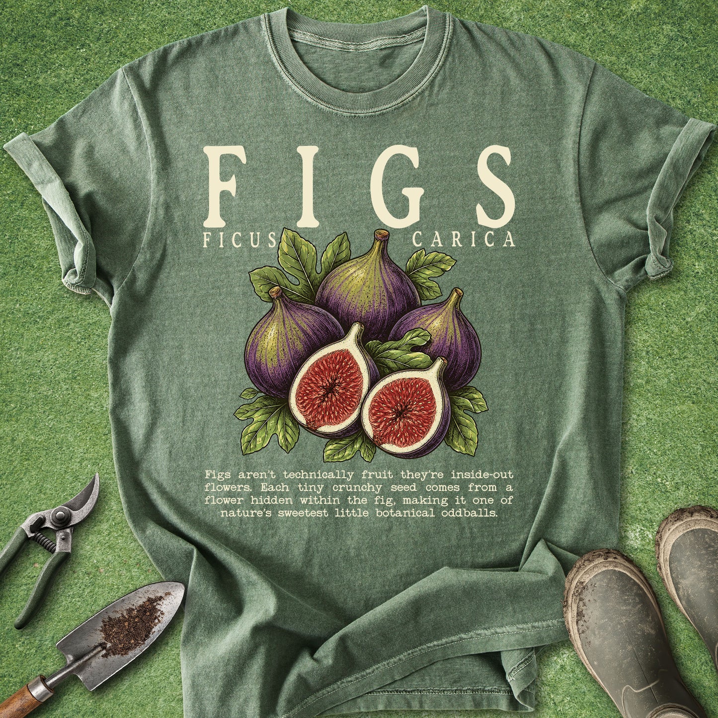 A sage green graphic t-shirt laid flat on a green grass-like surface, featuring an illustration of purple and red figs with leaves and the text 'FIGS FICUS CARICA' and a small gardening fact. Gardening props including pruning shears, a trowel, and shoes are placed around the shirt.