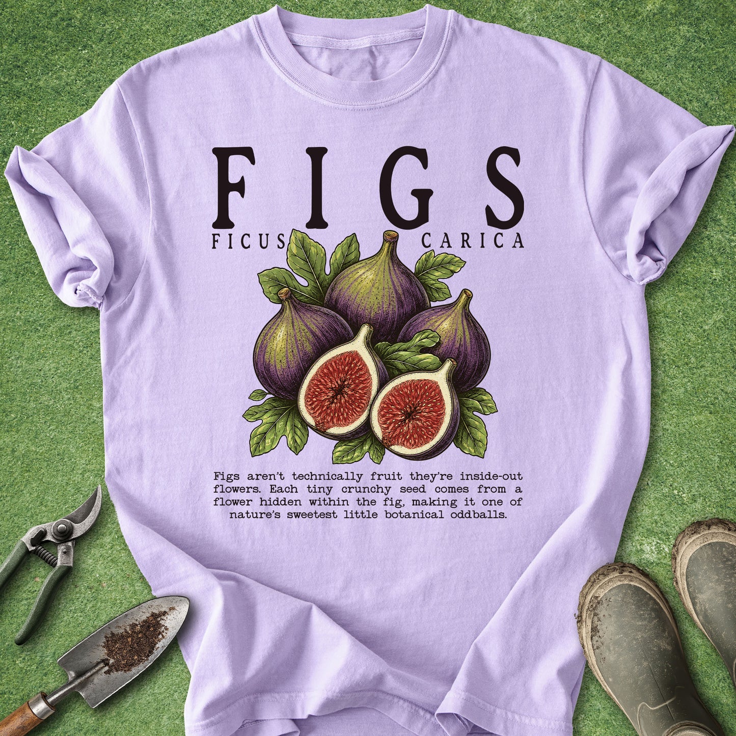 A light lavender t-shirt with a humorous 'Figs' botanical graphic, laid flat on a green grass background with gardening props like a trowel, pruning shears, and boots.