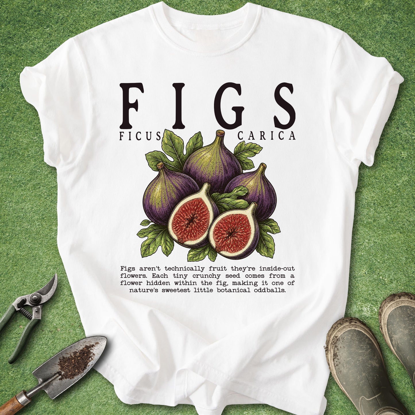 A white graphic t-shirt featuring a botanical illustration of figs and text, laid flat on a green grass-like surface with gardening props like pliers, a trowel, and boots.