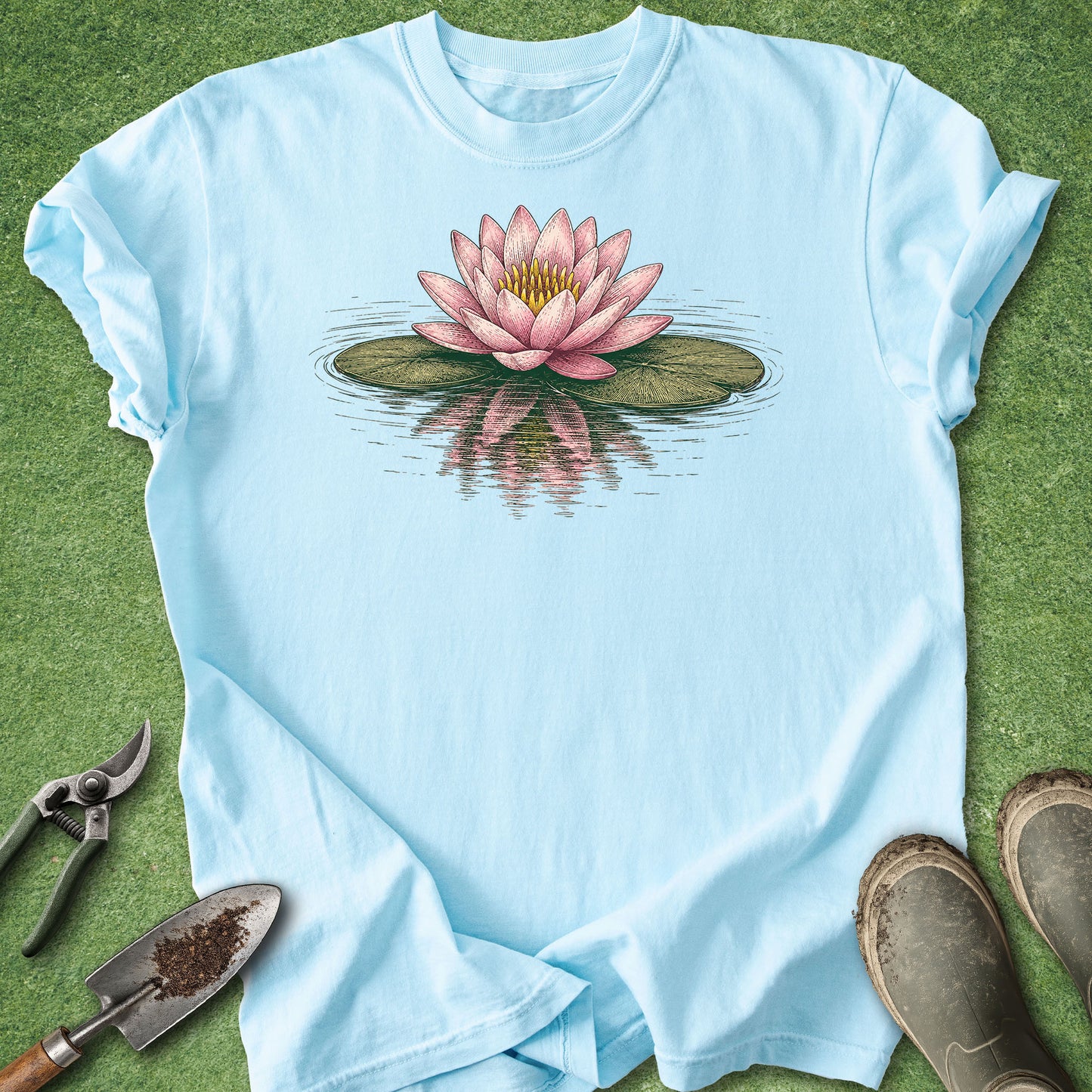 A light blue short-sleeve t-shirt laid flat on green grass, featuring a graphic of a pink water lily on a green lily pad with its reflection below. Gardening tools and muddy boots are styled around the shirt.