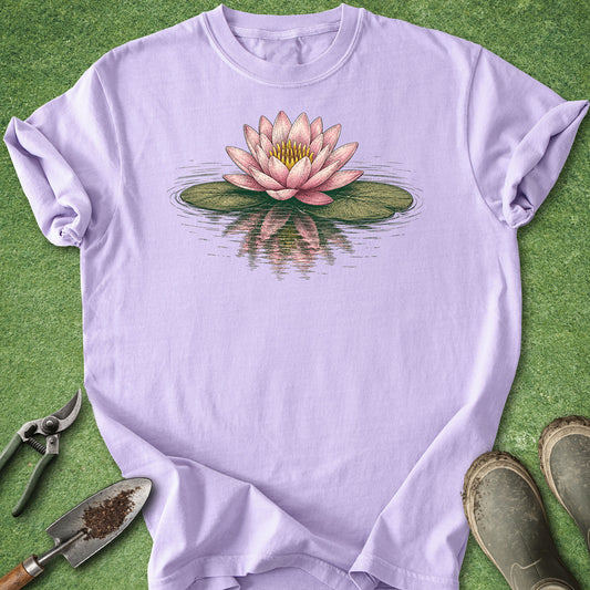 A light lavender crewneck t-shirt featuring a detailed graphic of a pink water lily on green lily pads with water reflection, laid flat on green grass with gardening tools and boots.