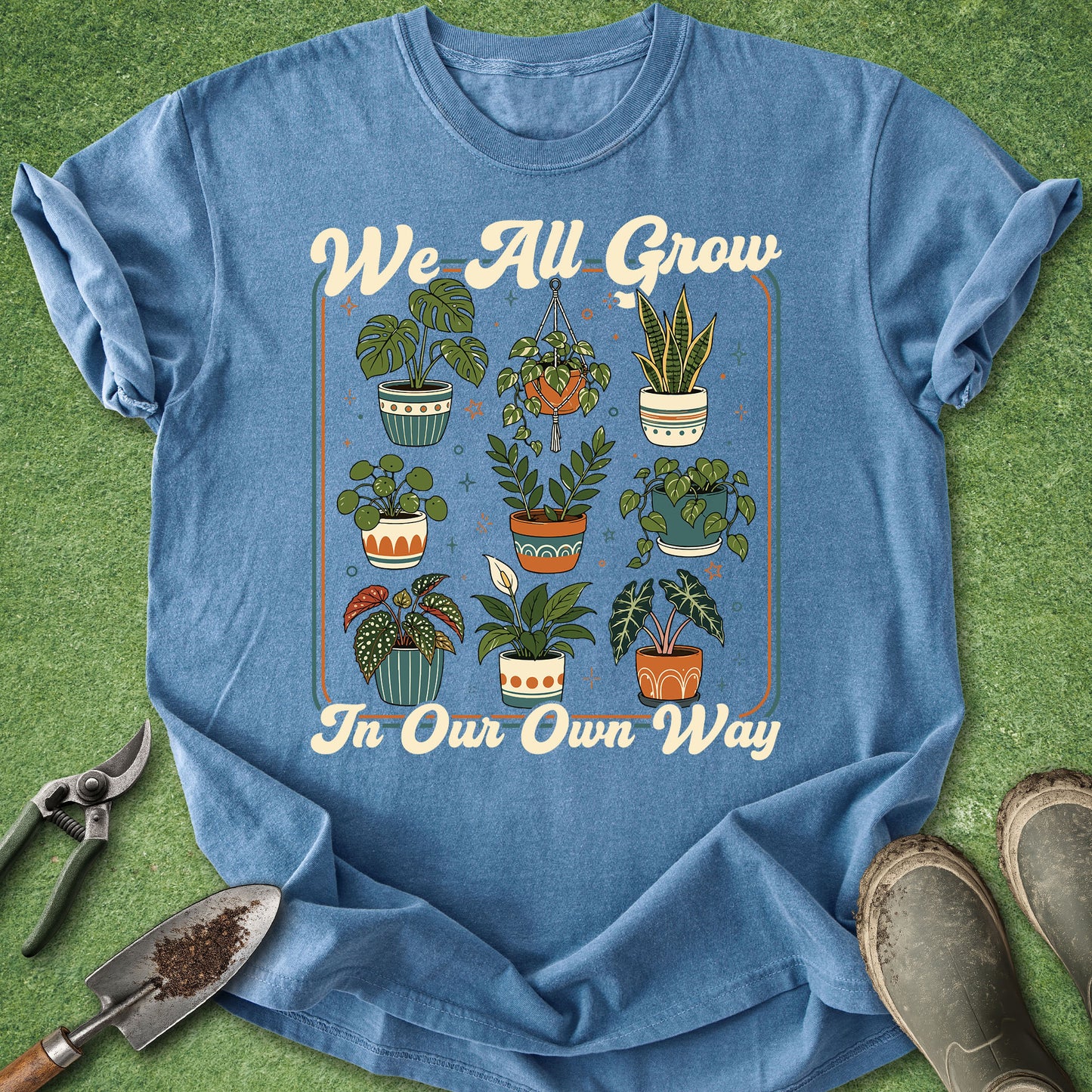 A blue graphic t-shirt laid flat on green grass-like turf, surrounded by gardening props including pruning shears, a trowel, and muddy boots. The shirt features a retro-style illustration of various potted houseplants with the text 'We All Grow In Our Own Way' in cream lettering.