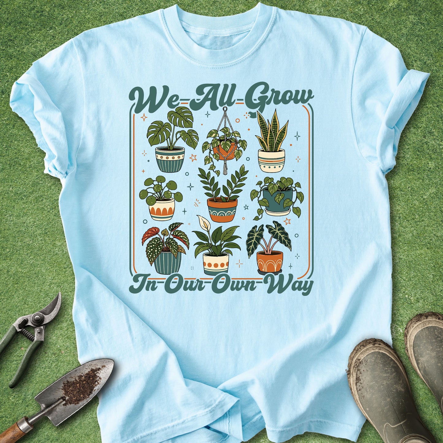 A light blue graphic t-shirt laid flat on green grass, featuring a retro-style illustration of various potted houseplants with the text 'We-All-Grow' at the top and 'In-Our-Own-Way' at the bottom. Gardening props, including pruning shears, a trowel, and boots, are arranged around the shirt.