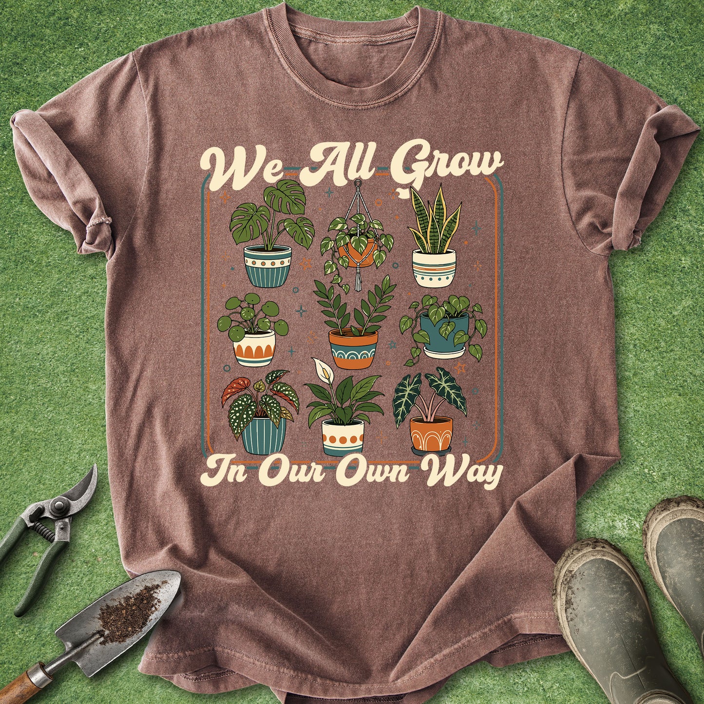 A heathered brown graphic t-shirt laid flat on green grass-like artificial turf, with gardening tools (shears, trowel) and boots as props. The shirt features a central graphic of various potted plants and the text 'We All Grow In Our Own Way'.