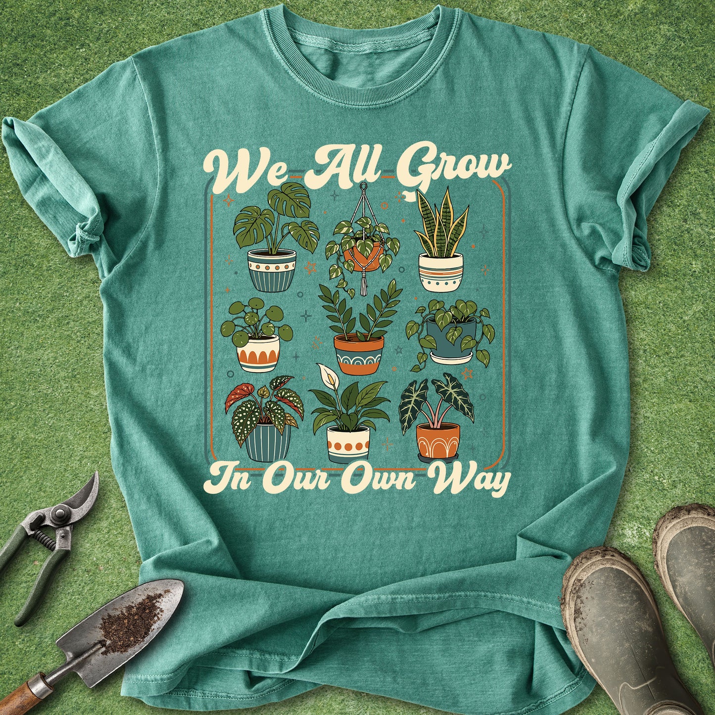 A teal graphic t-shirt laid flat on a green grass-like surface, featuring the phrase 'We All Grow In Our Own Way' around an illustration of potted plants. Gardening props including pruning shears, a trowel, and boots are placed around the shirt.
