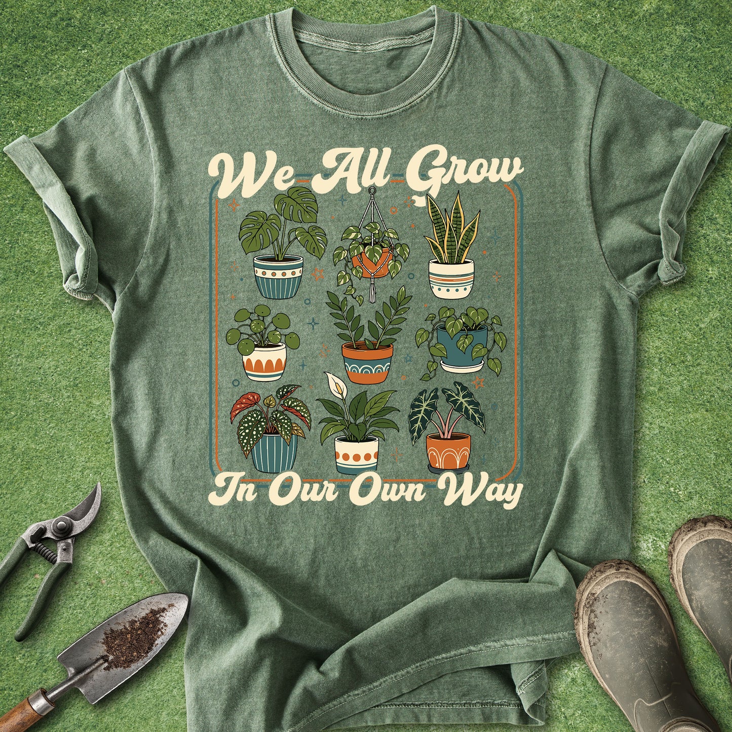 A sage green graphic t-shirt laid flat on a textured green grass background, styled with gardening tools (trowel, pruning shears) and muddy boots. The t-shirt features a retro-style illustration of various potted plants in a central frame, with the cream-colored text 'We All Grow' above and 'In Our Own Way' below.