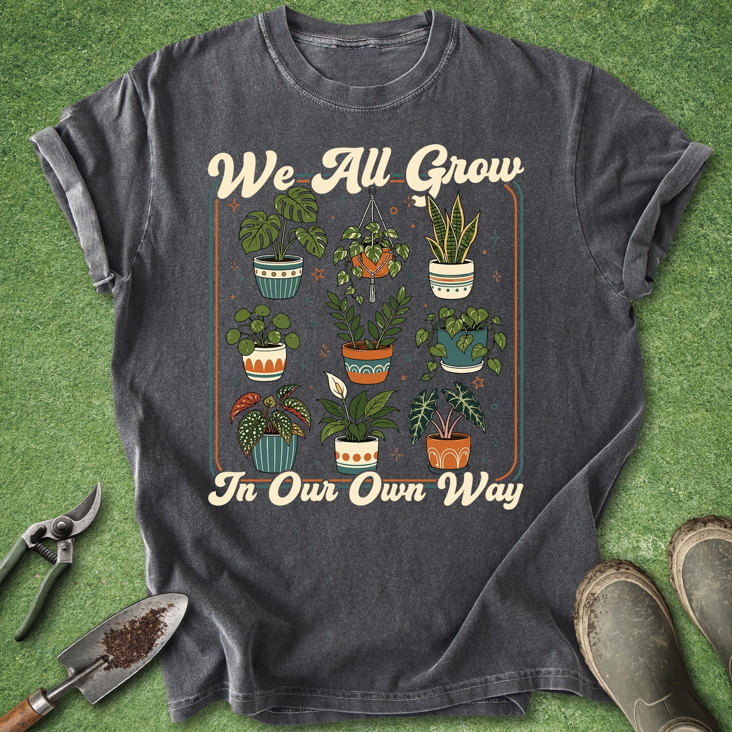 A dark gray graphic t-shirt laid flat on a green grass-like surface, surrounded by gardening props including pruning shears, a trowel, and boots. The shirt features a retro-style print with the text 'We All Grow In Our Own Way' and an illustration of various potted plants.