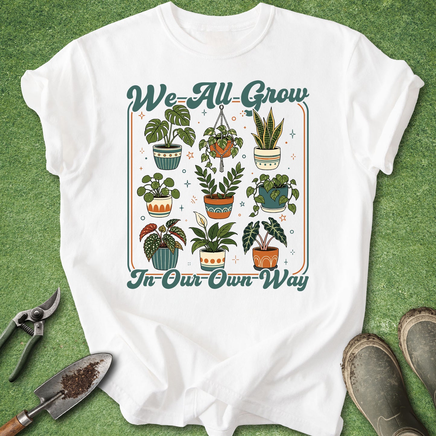 A white graphic t-shirt laid flat on a green grass-like surface, featuring the phrase 'We All Grow In Our Own Way' around an illustrated arrangement of potted plants. Gardening tools and boots are placed around the shirt as styling props.