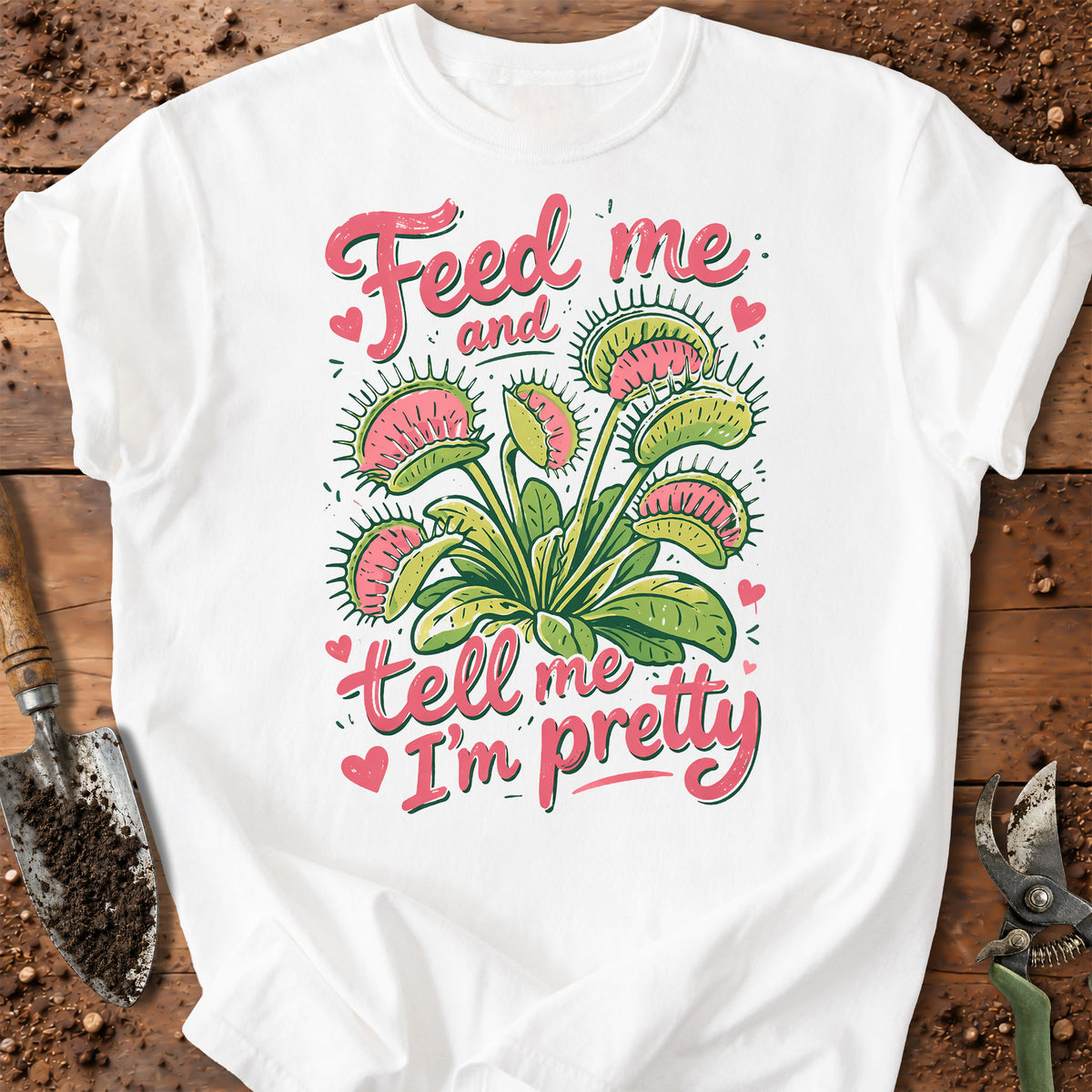 A white short-sleeve T-shirt laid flat on a rustic wooden surface with gardening props. The shirt features a colorful graphic of a Venus flytrap plant in green and pink, surrounded by the phrase 'Feed me and tell me I'm pretty' in pink script, with small pink hearts.