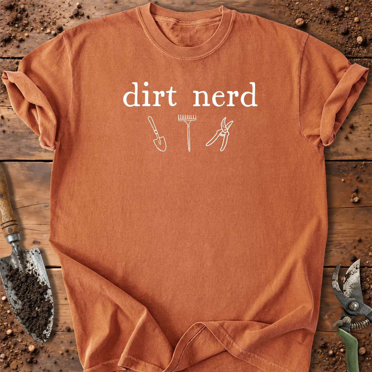 An orange crewneck t-shirt with a white graphic that reads 'dirt nerd' above simple line drawings of gardening tools (a trowel, a rake, and pruning shears), laid flat on a rustic wooden background with gardening props.