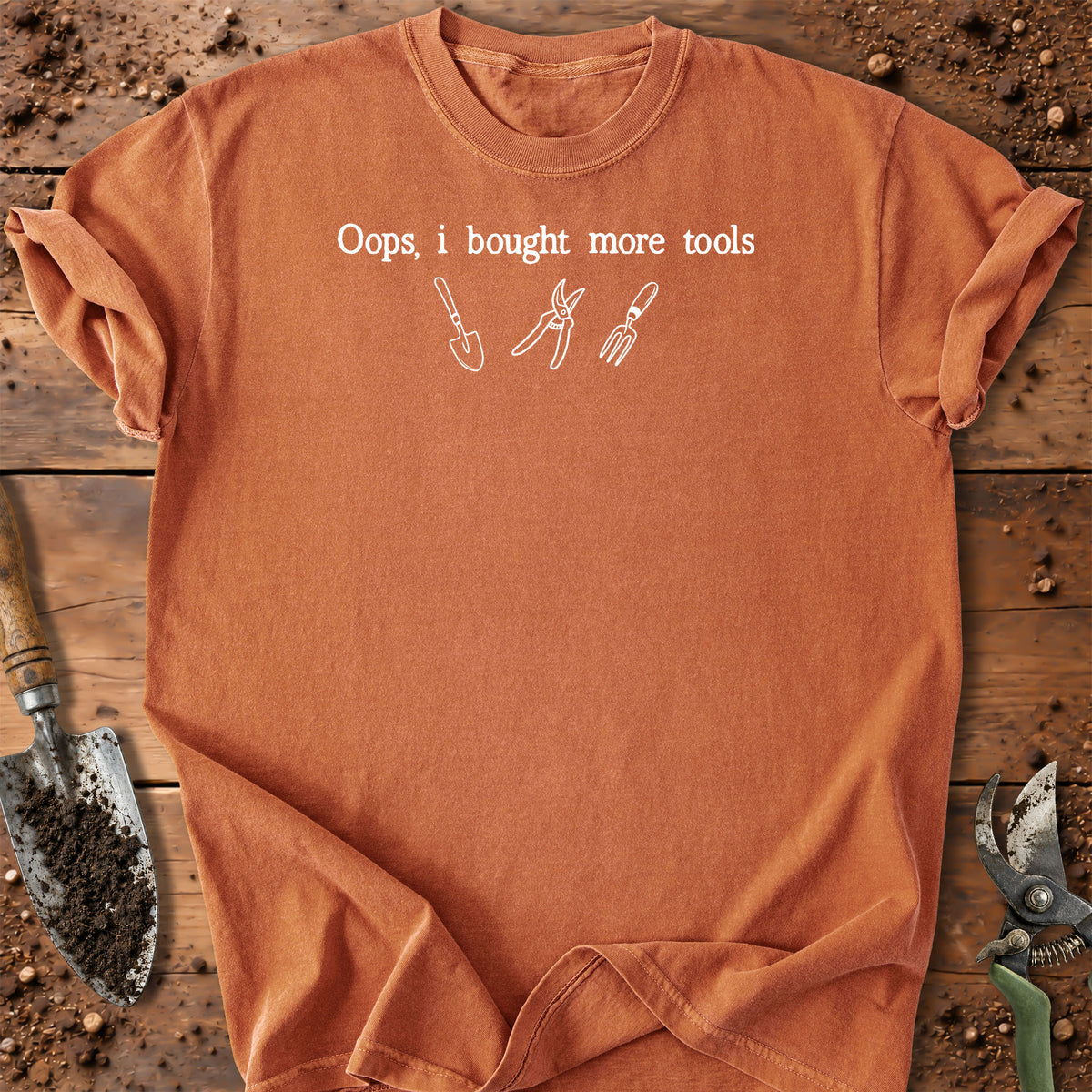An orange short-sleeve crewneck t-shirt with a white graphic print reading 'Oops, i bought more tools' above simple line drawings of gardening tools. The shirt is laid flat on a rustic wooden surface surrounded by gardening props like a trowel and pruning shears.