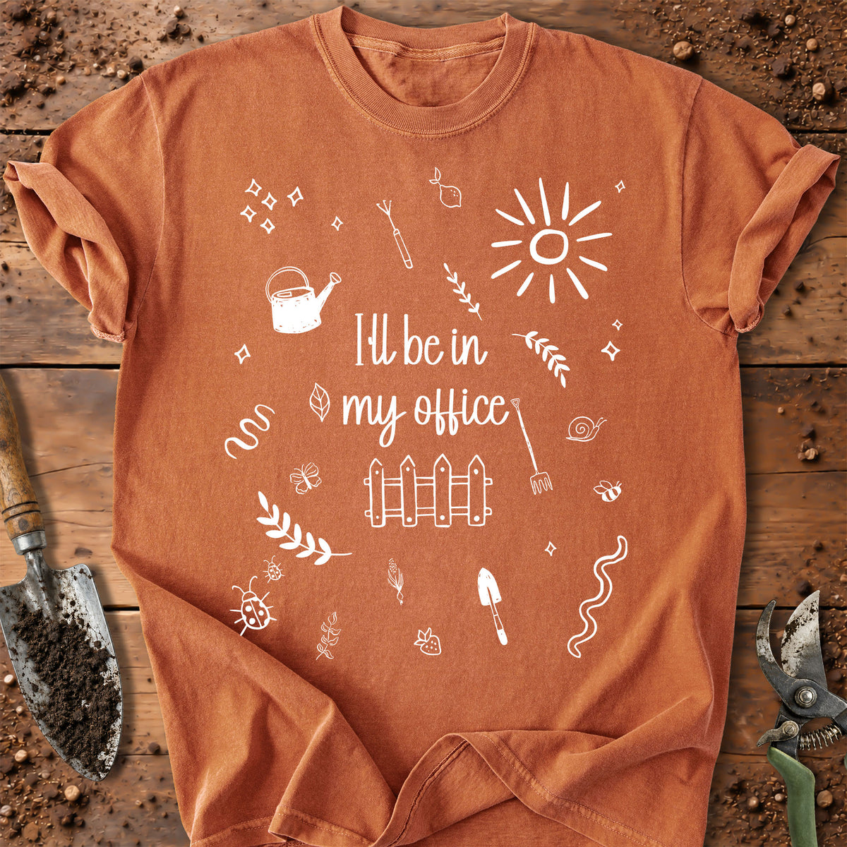 An orange short-sleeve t-shirt with a white graphic print that reads 'I'll be in my office' surrounded by gardening doodles, laid flat on a rustic wooden background with gardening tools.