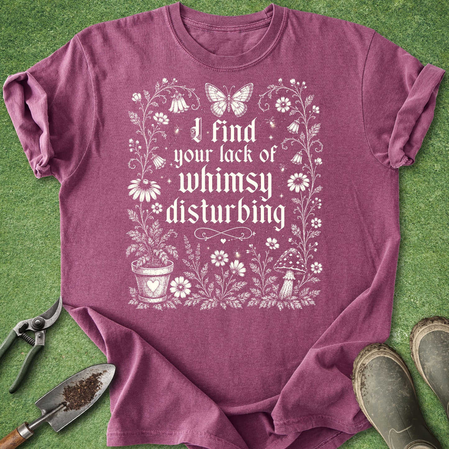 A heathered purple short-sleeve t-shirt with a white graphic print that reads 'I find your lack of whimsy disturbing' surrounded by flowers, butterflies, and a potted plant. The shirt is laid flat on green grass with gardening tools (trowel, pruning shears) and muddy boots nearby.
