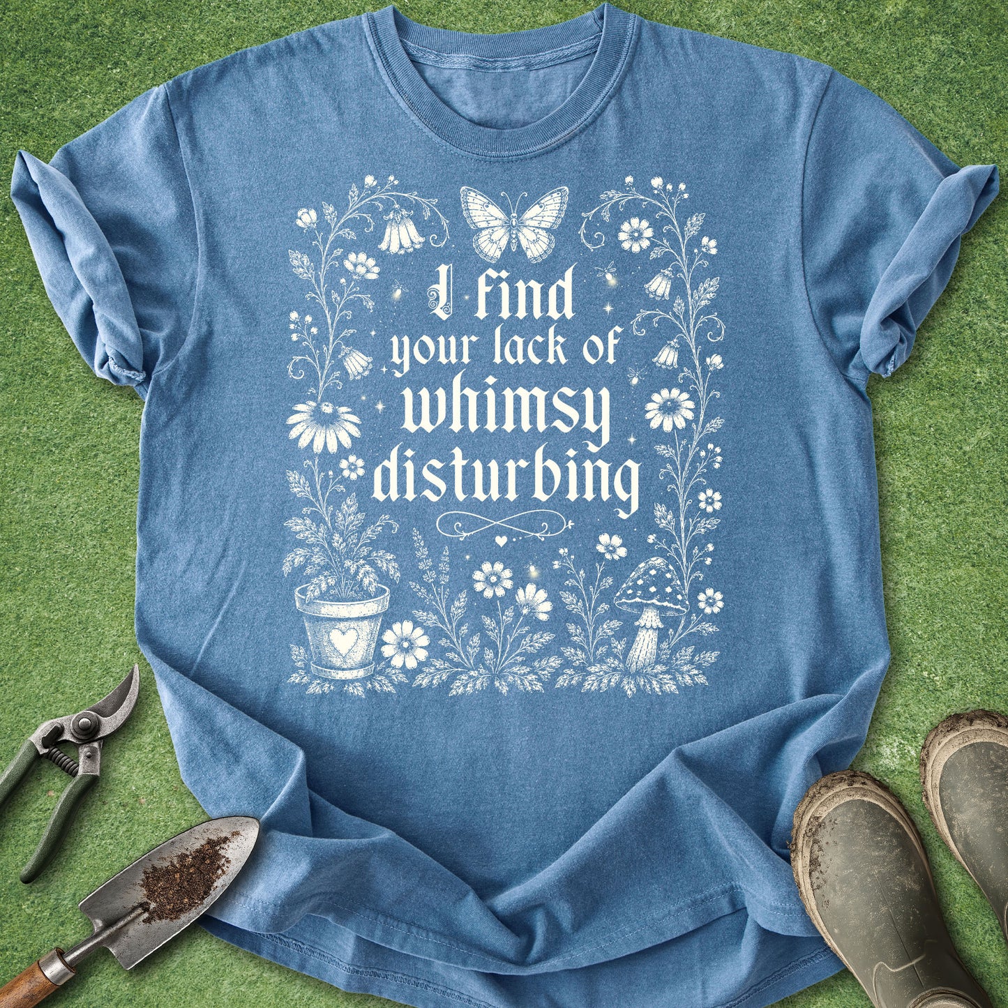 A blue short-sleeve graphic T-shirt laid flat on green grass-like background, with gardening tools and boots around it. The shirt has a white floral and butterfly print with the text, 'I find your lack of whimsy disturbing.