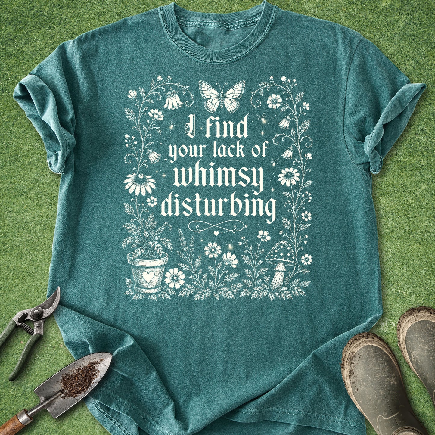 A teal short-sleeve graphic T-shirt laid flat on green grass with gardening props around it. The shirt has a white floral and butterfly print with the text 'I find your lack of whimsy disturbing.
