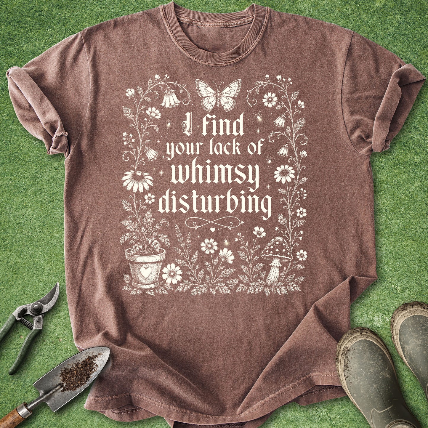 A brown short-sleeve graphic t-shirt laid flat on green grass-like background, surrounded by gardening props including pruning shears, a trowel, and boots. The shirt features a white floral and butterfly design with the text 'I find your lack of whimsy disturbing'.