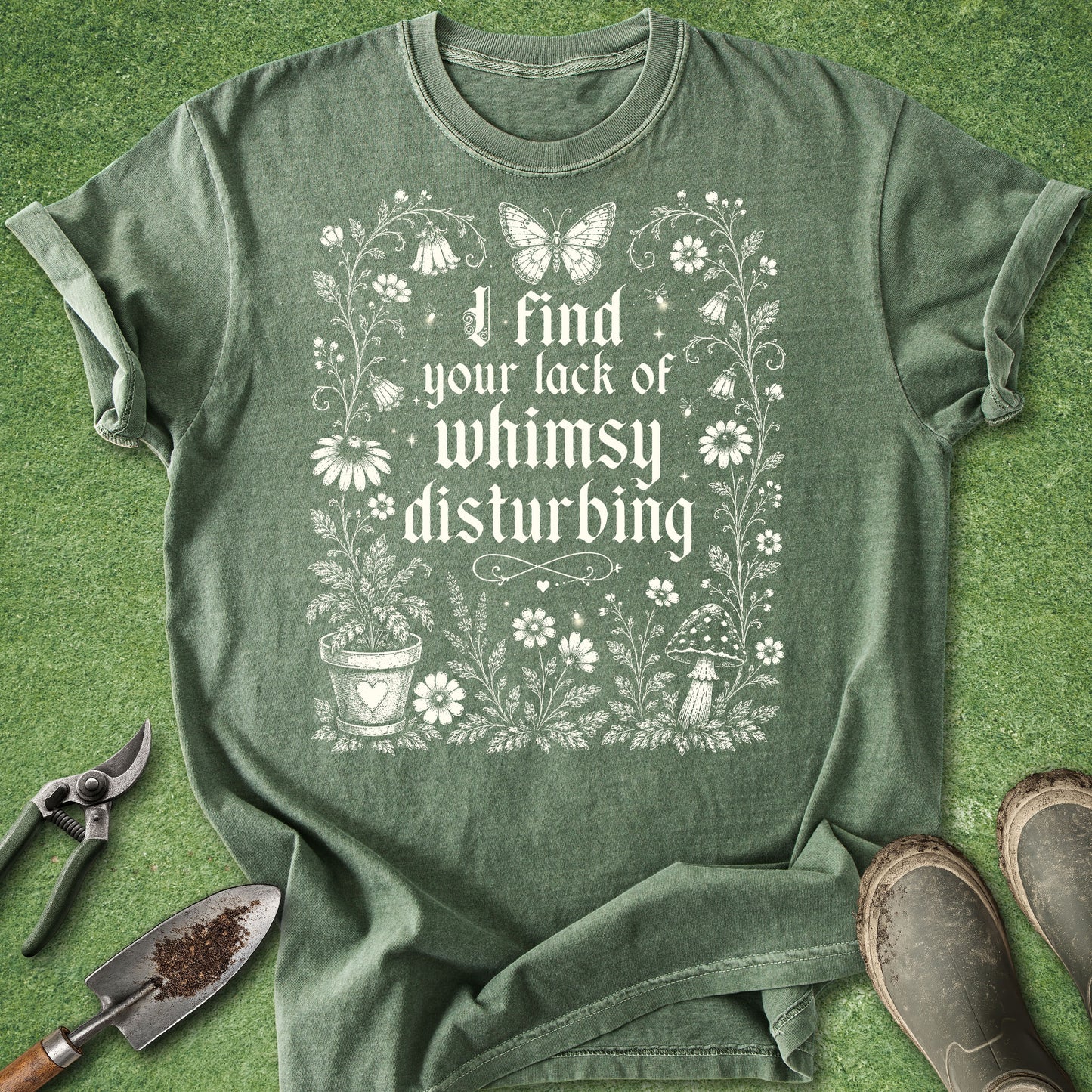 A sage green graphic t-shirt laid flat on a green grass-like surface, surrounded by gardening props including pruning shears, a trowel, and boots. The shirt features a white distressed graphic with a butterfly, flowers, a potted plant, and the text 'I find your lack of whimsy disturbing'.